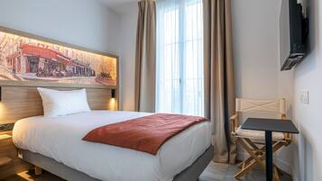 Ein Hotelzimmer mit Bett, Sessel, Tisch, Fernseher und einem großen Fenster mit Vorhängen.