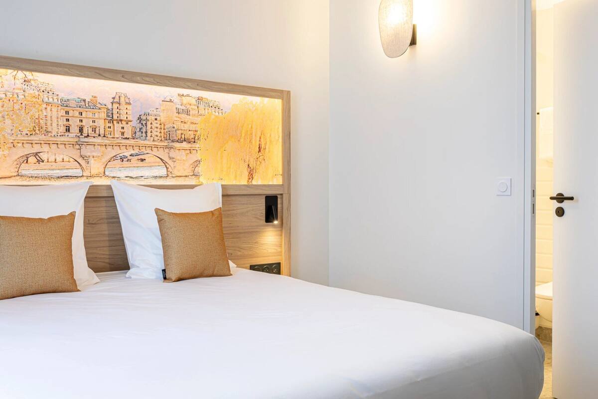 A neatly made bed with white linens and two pillows, a wall-mounted light fixture, and a door with a handle.