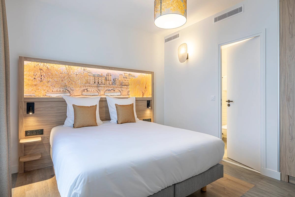 A hotel room with a large bed, a cityscape mural on the wall, a door, and a nightstand with a lamp.