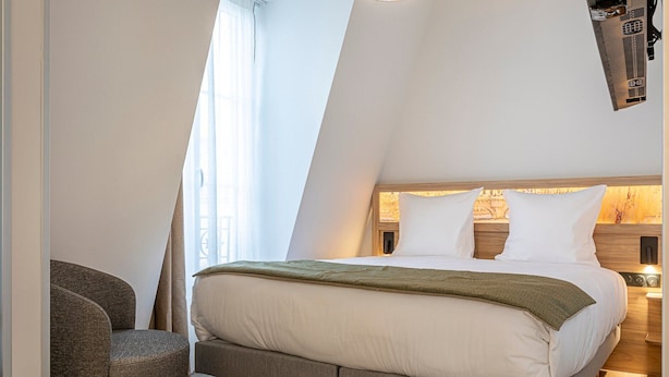 Een moderne slaapkamer met een schuine plafond, een bed met witte lakens, een groene plaid en een houten nachtkastje met een lamp.
