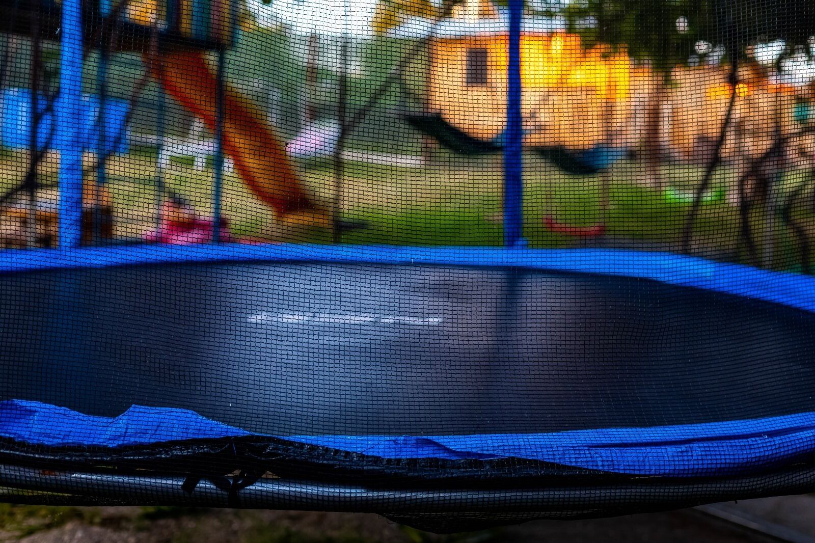 Een trampoline met een blauw veiligheidsnet, tegen een achtergrond van een gazon en een houten speelconstructie.