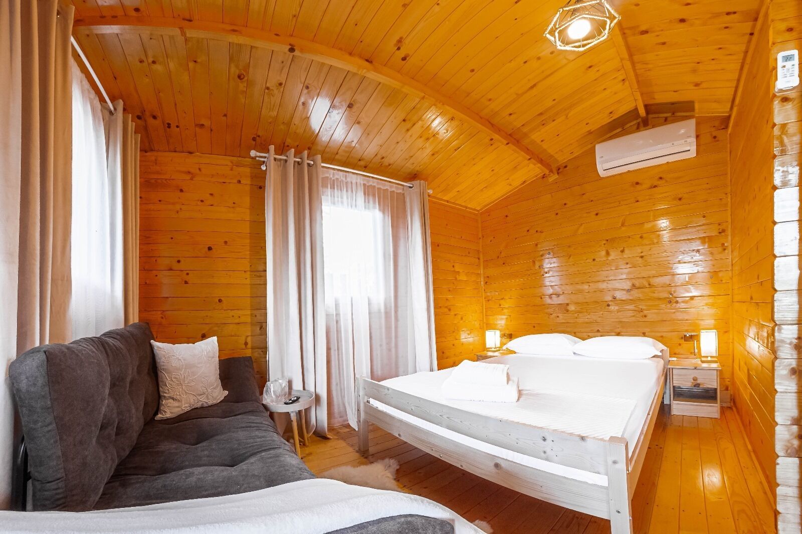 A cozy cabin room with a wooden ceiling, a bed with white linens, a gray sofa, and a small table with a lamp.