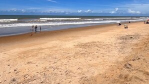 A wide sandy beach with gentle waves, clear skies, and a few people and beach chairs.