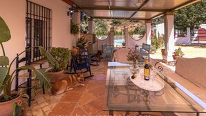 A covered patio with a glass table set for dining, surrounded by potted plants and outdoor furniture.