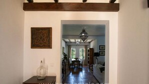 A hallway with a wooden beam ceiling, a chandelier, a dining area with a table and chairs, and a sofa in the background.