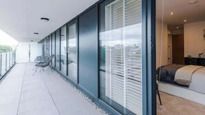 A balcony with a chair, a bed with a headboard, and a window with blinds. - Luxury City Apt with Balcony & Parking (Leeds)