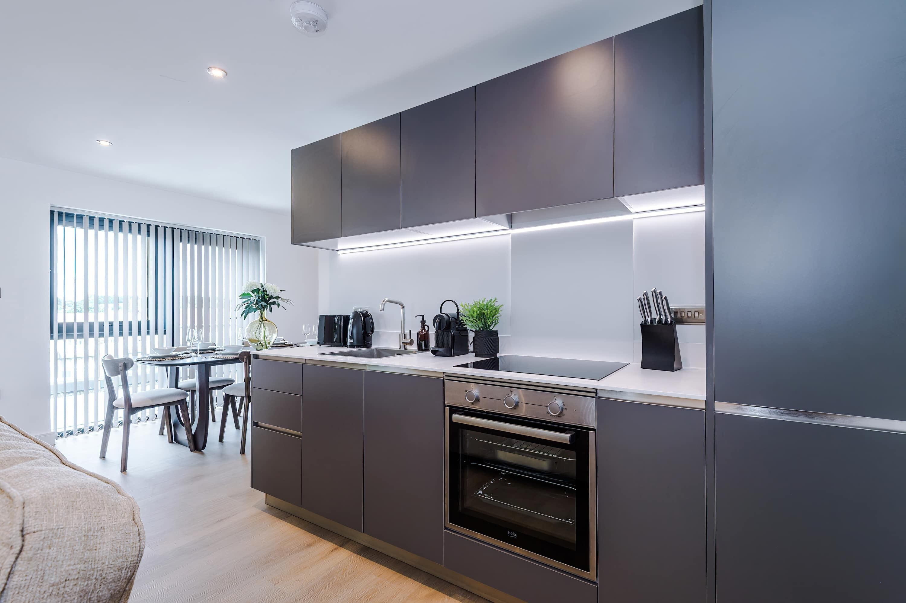 A modern kitchen with dark cabinetry, a built-in oven, and a dining area with a table and chairs.