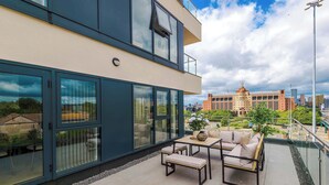 Un bâtiment moderne doté de grandes fenêtres, d’un balcon avec mobilier d’extérieur et offrant une vue sur le paysage urbain.