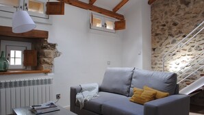 Un salon avec un canapé gris, des poutres en bois, un mur en pierre et un escalier.