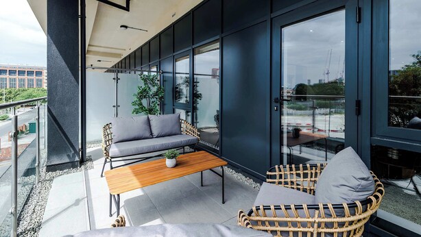 En modern utomhusplats med sittmöbler, bestående av en soffa, en fåtölj i rotting och ett träbord, med utsikt över en byggarbetsplats.