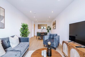 A modern living room with a grey sofa, a blue armchair, a coffee table with decorative items, a TV on a wooden stand, and a dining area in the background. - Chic City 2 Bed Apt with Terrace & Parking (Leeds)