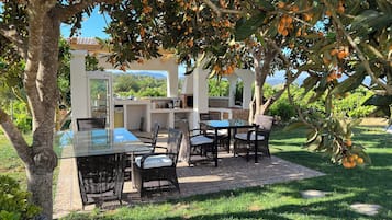 Een buitenterras met een glazen tafel, rieten stoelen, een barbecue en uitzicht op de omliggende begroeiing.