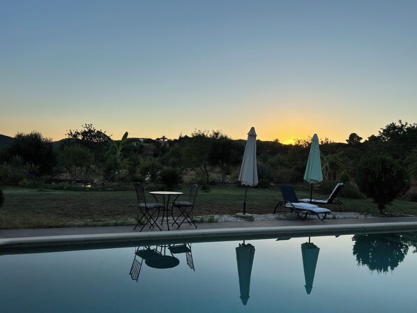 A poolside area with lounge chairs and umbrellas, surrounded by greenery and a clear sky.