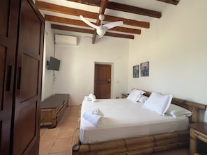 Quarto com cama de madeira, um ventilador de teto, TV fixada na parede e uma porta.