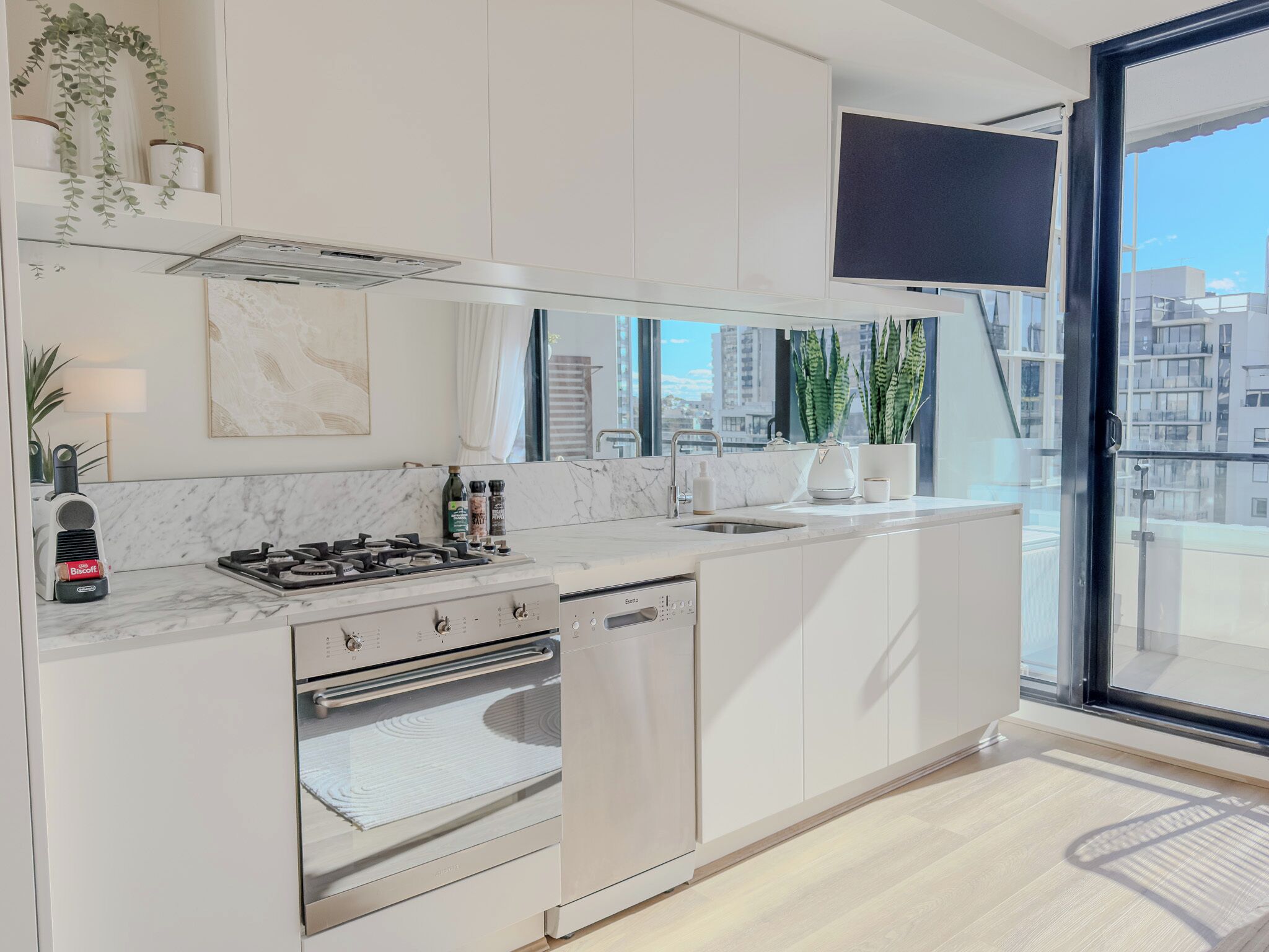 A modern kitchen with white cabinetry, a built-in oven, and a stove. There are potted plants on the windowsill and a large window offering a view of buildings.
