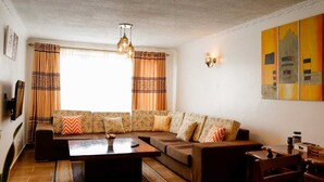 Een woonkamer met een modulaire bank, een salontafel en een televisie. Gordijnen, een wandlamp en een schilderij zijn aanwezig.