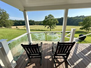 Um deck coberto com duas cadeiras de madeira e uma mesa, com vista para um campo gramado e um lago.