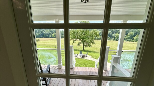 Vue de l’intérieur d’une maison, à travers une grande fenêtre, sur une terrasse avec une table et des chaises, un arbre et une pelouse.