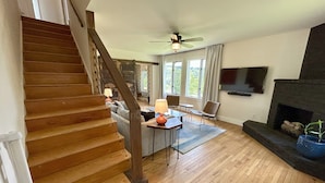 Un vaste espace de vie agrémenté d’un escalier en bois, d’une cheminée, d’un téléviseur fixé au mur et d’un coin repas avec une table et des chaises.