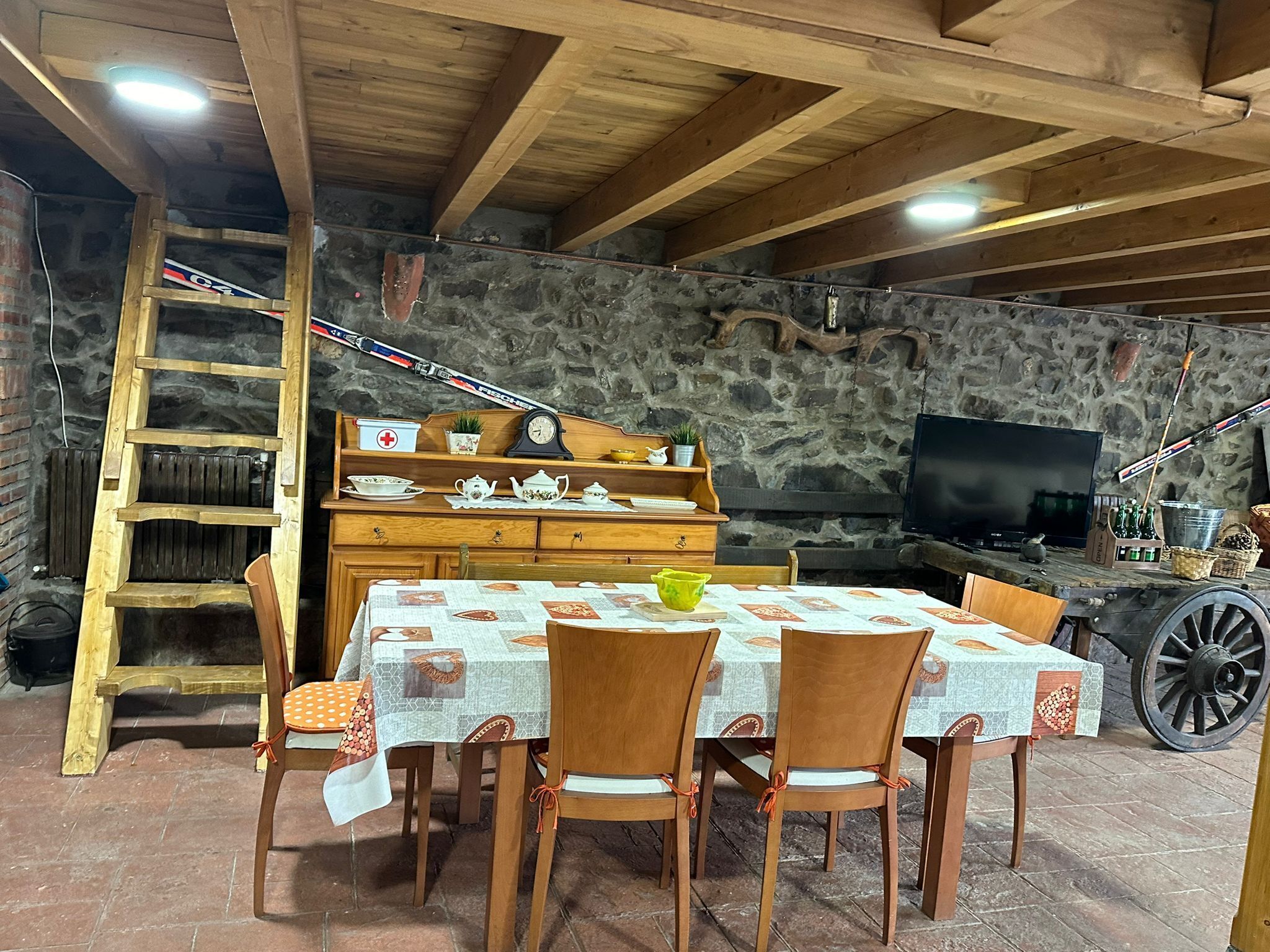 Een rustieke eethoek met een houten tafel voor vier personen, omringd door stoelen. Een houten ladder en een stenen muur zijn zichtbaar op de achtergrond.