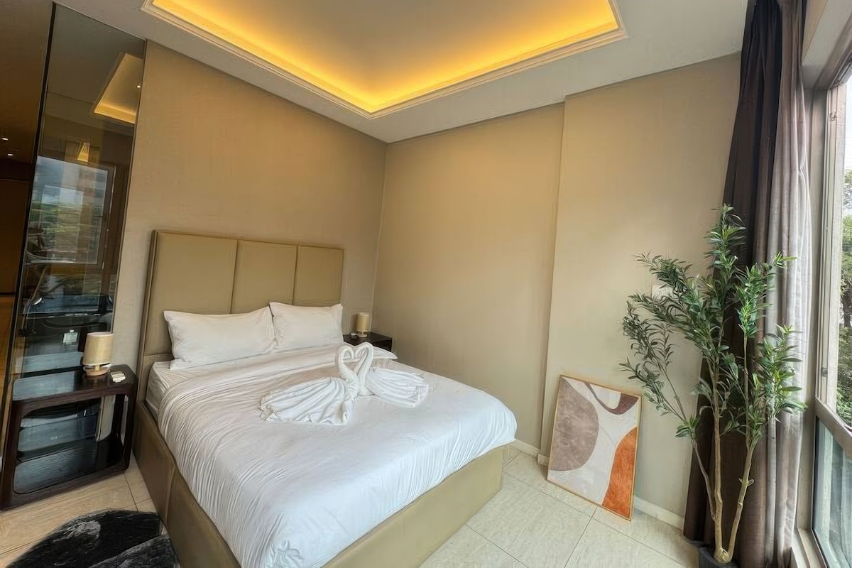 Habitación de hotel con cama, mesitas de noche, un espejo, ventana con cortinas y una planta en maceta.
