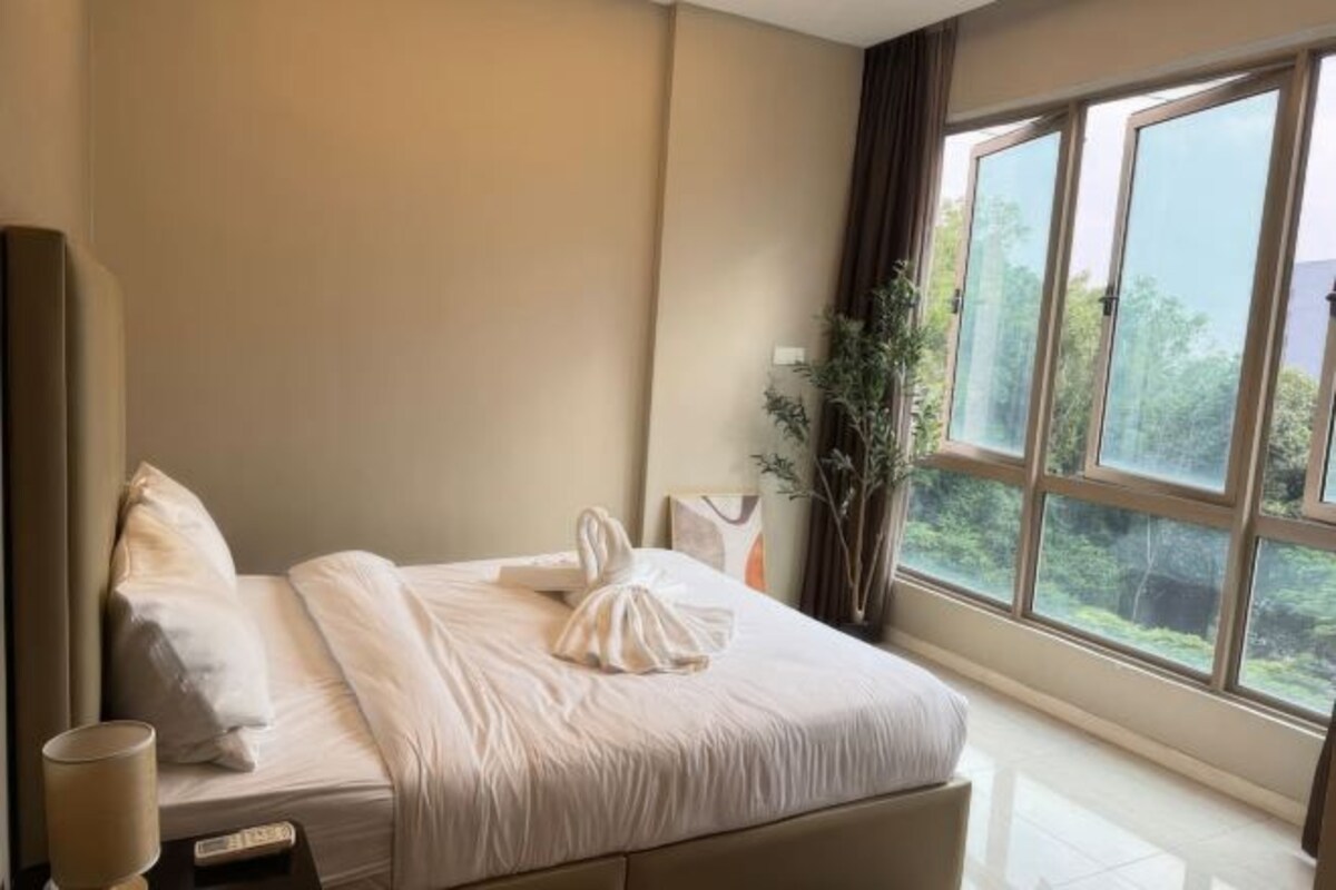 Una habitación de hotel con un ventanal grande, una cama con ropa blanca, una mesita de noche con lámpara y una planta.