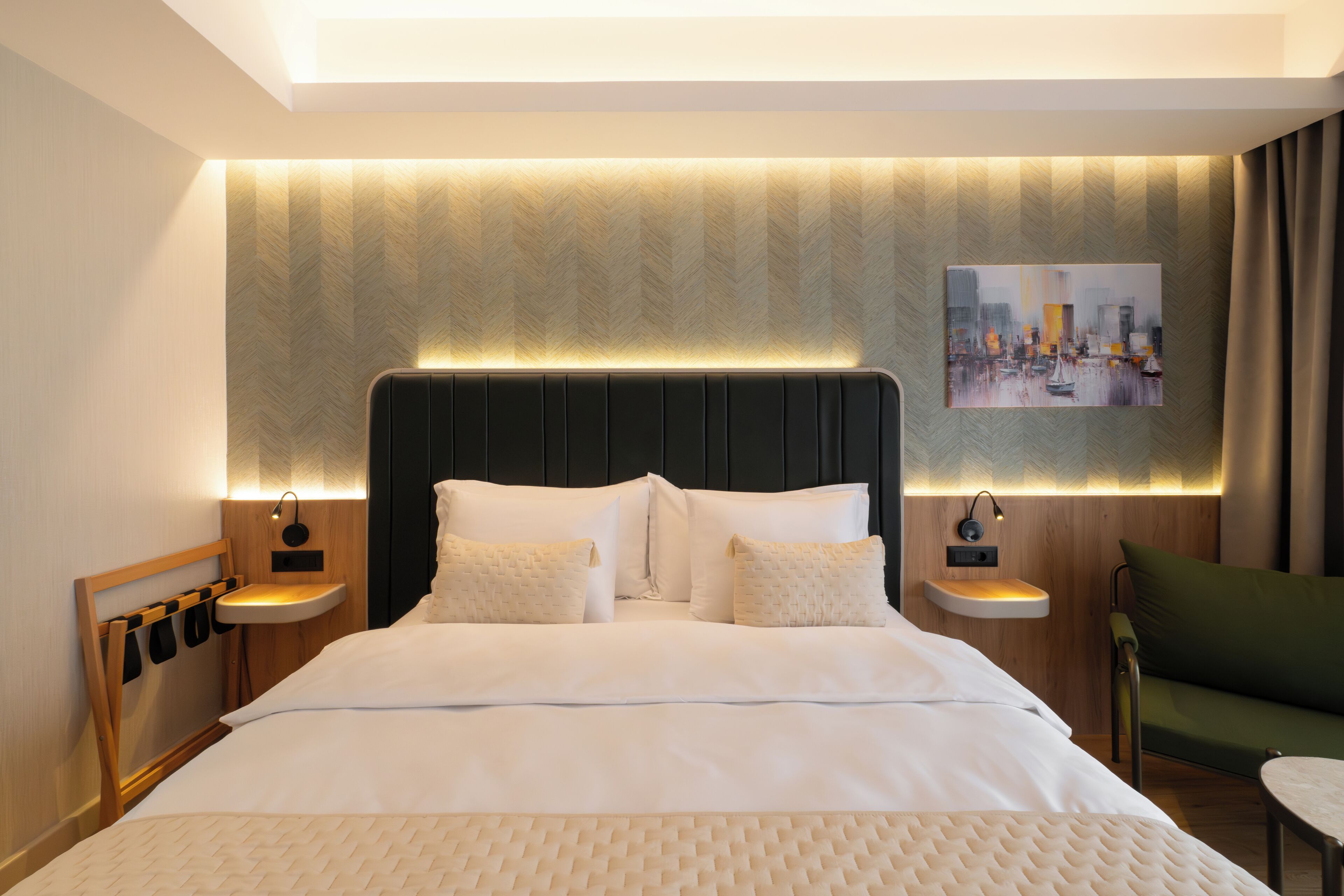 A modern hotel room with a large bed, a nightstand with a lamp, a green armchair, and a painting on the wall.