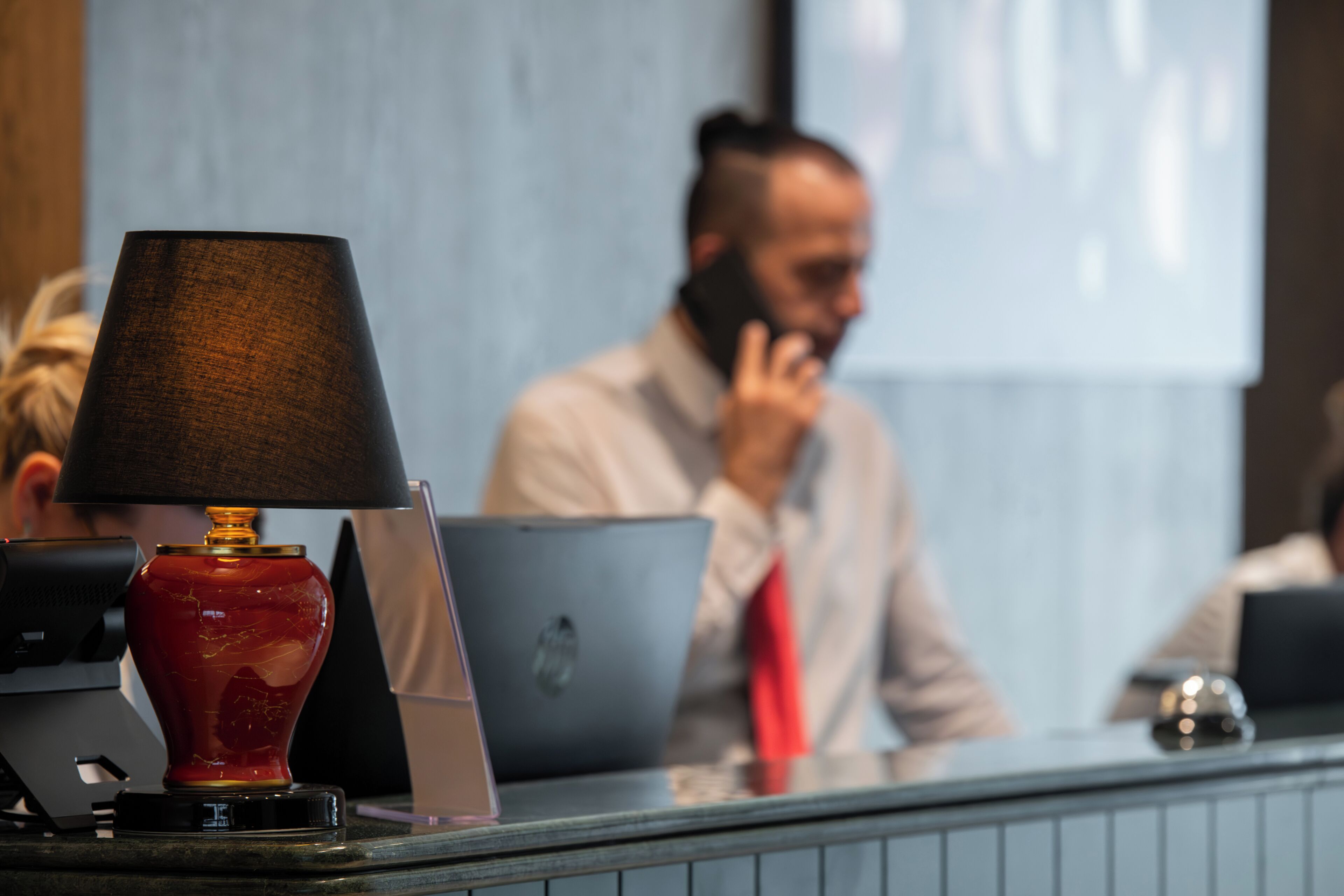 A reception desk with a red vase lamp, laptops, and a blurred background of a person on the phone.