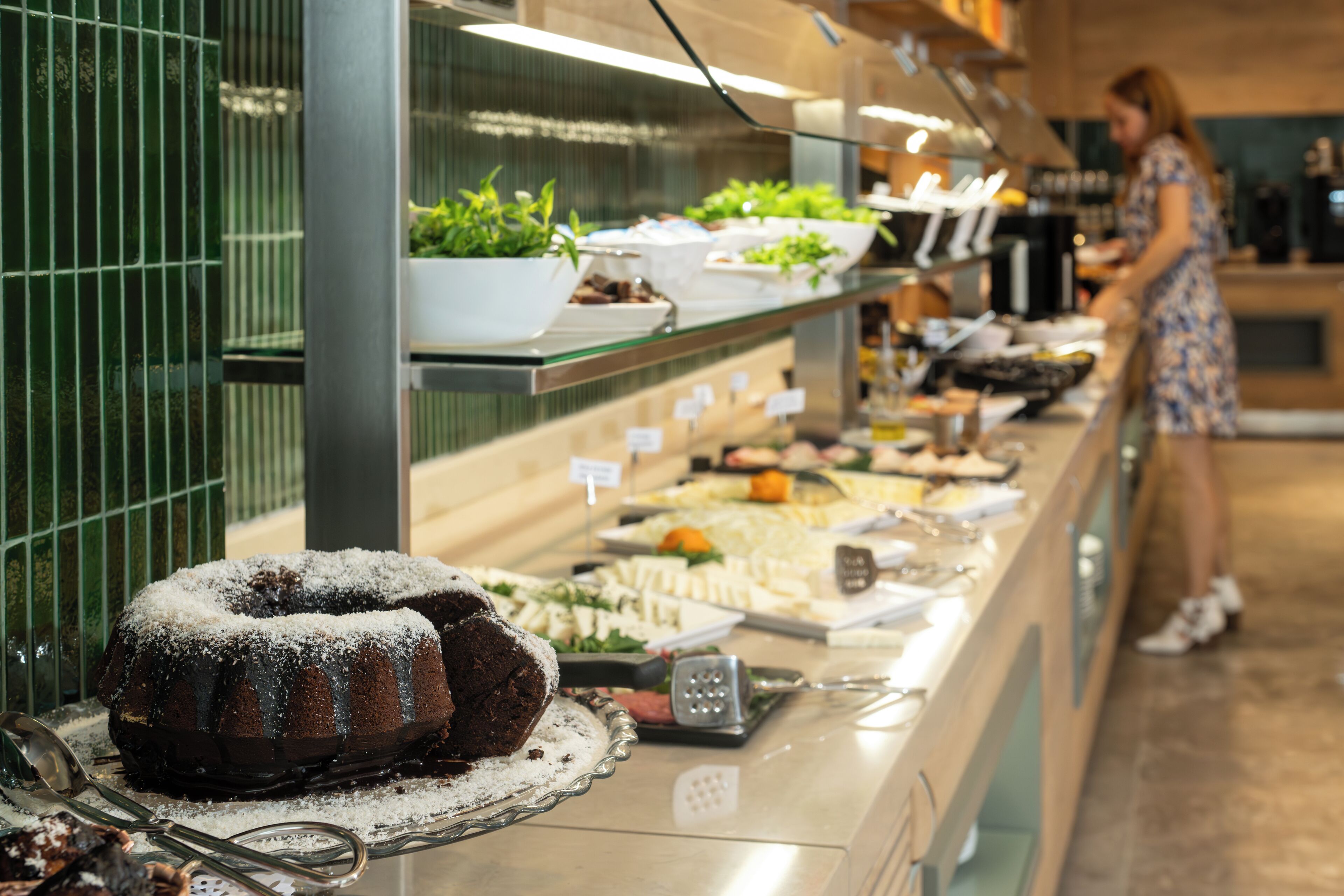 A buffet with various food items, including a chocolate bundt cake, and a woman serving food in the background.