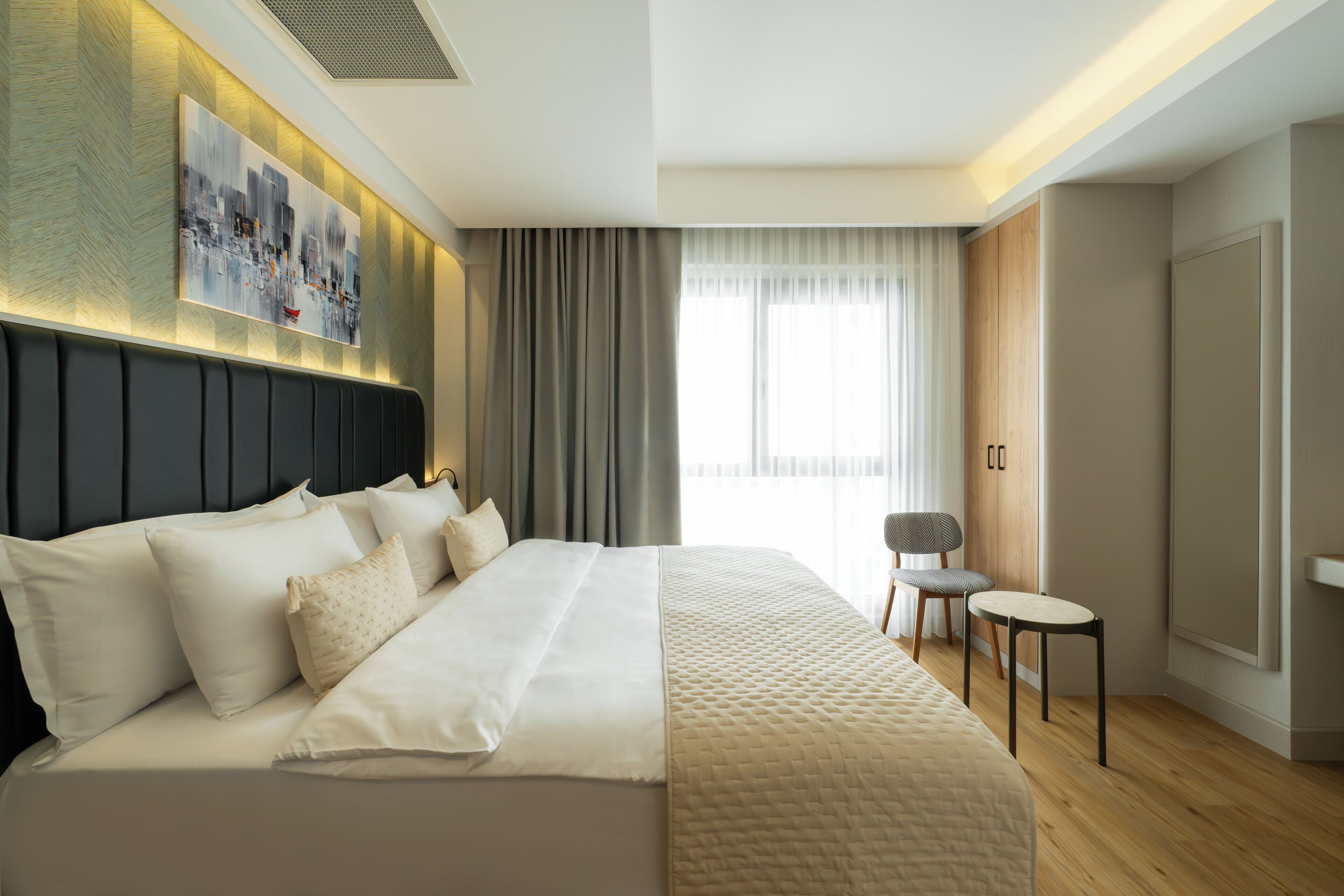 Ein modernes Hotelzimmer mit einem großen Bett, einer Sitzecke inklusive Sessel und Tisch, einem Bild an der Wand und einem großen Fenster mit Vorhängen.