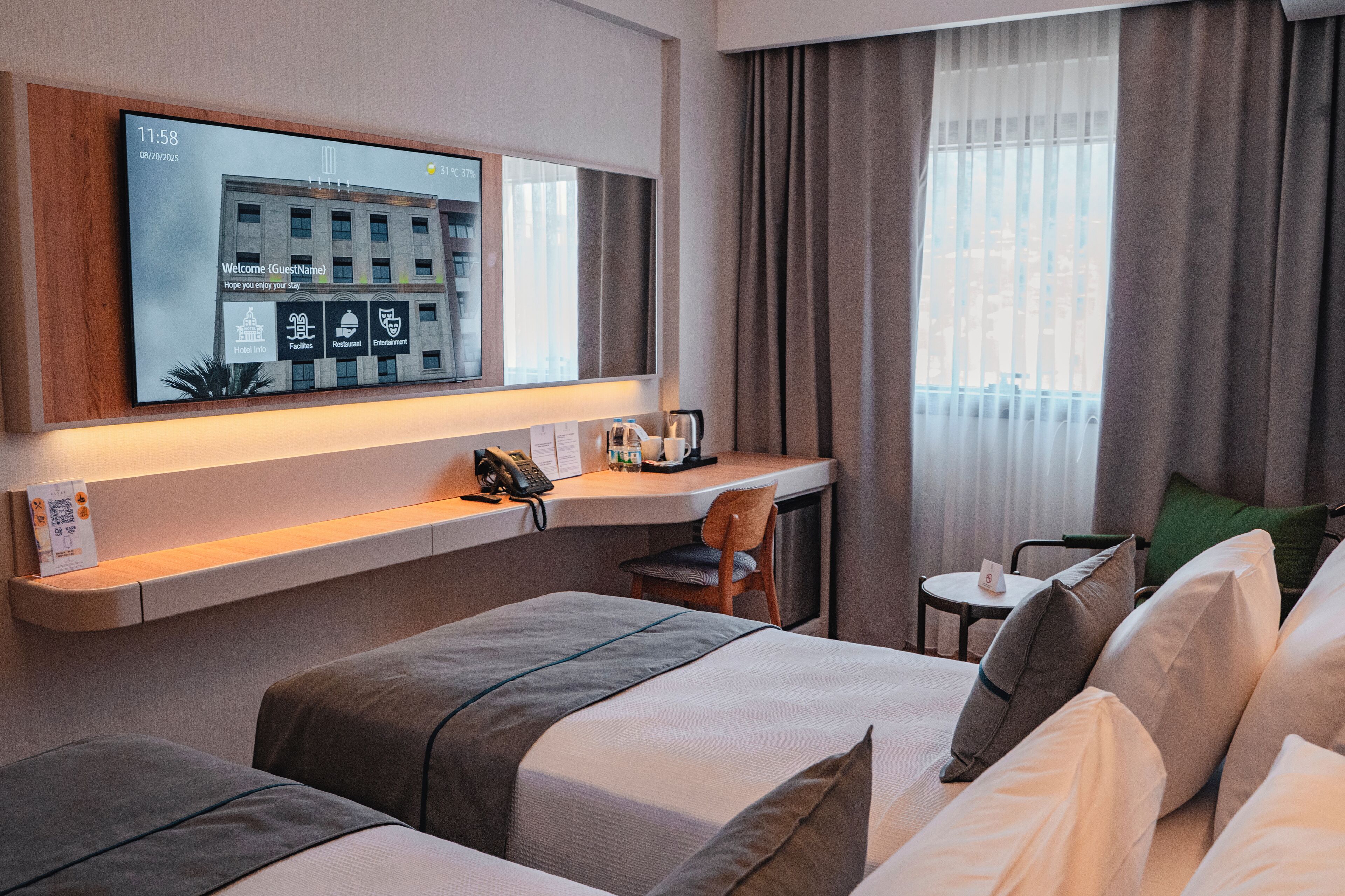 A hotel room with a bed, a desk, a chair, a TV mounted on the wall, and a window with curtains.