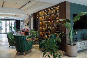 Un hall con sillas verdes, una mesa de centro, una planta grande en maceta y una pared con estantes decorativos.