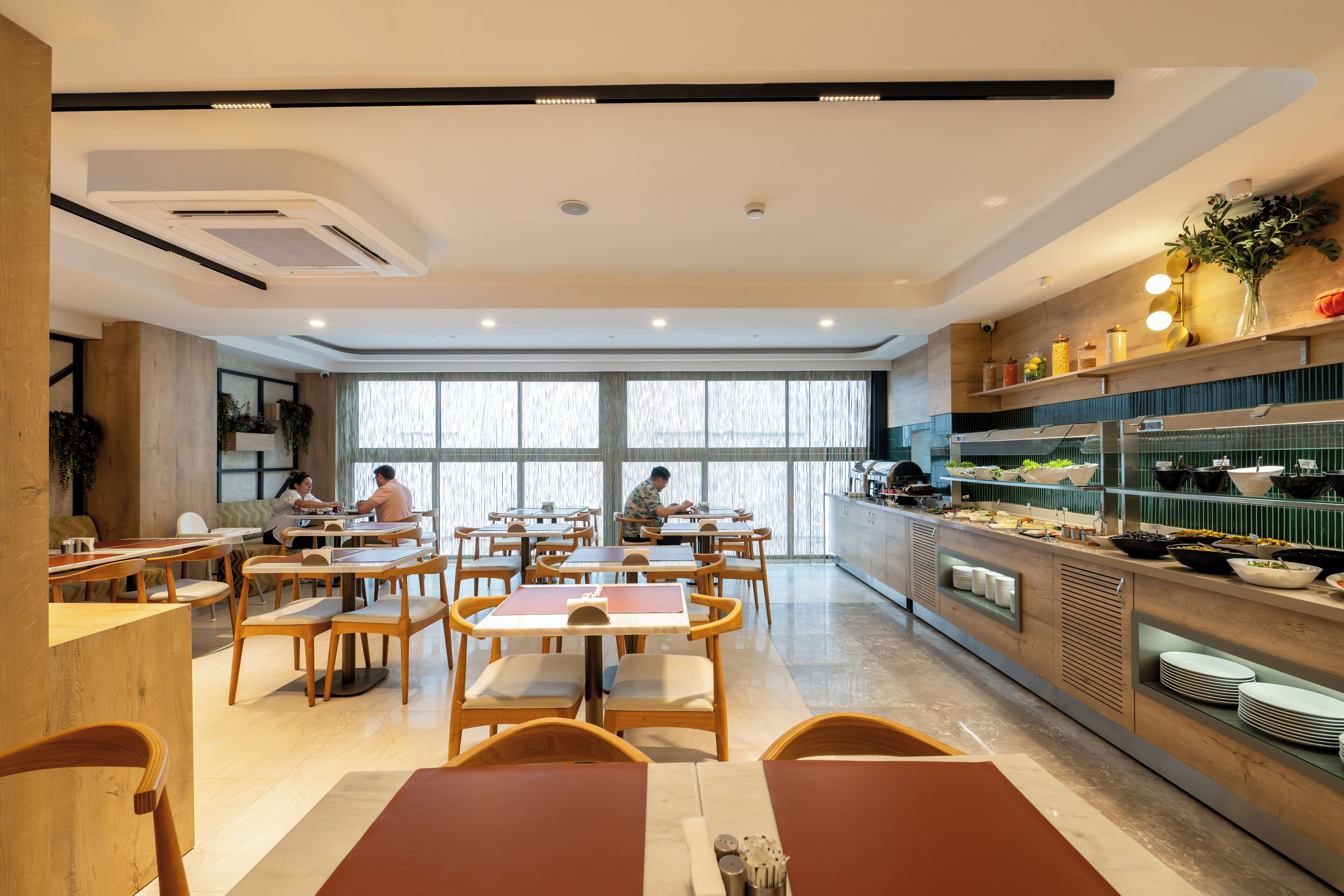 Uma sala de jantar moderna com mesas e cadeiras de madeira, um balcão de buffet com diversos pratos, e janelas amplas que permitem entrada de luz natural.