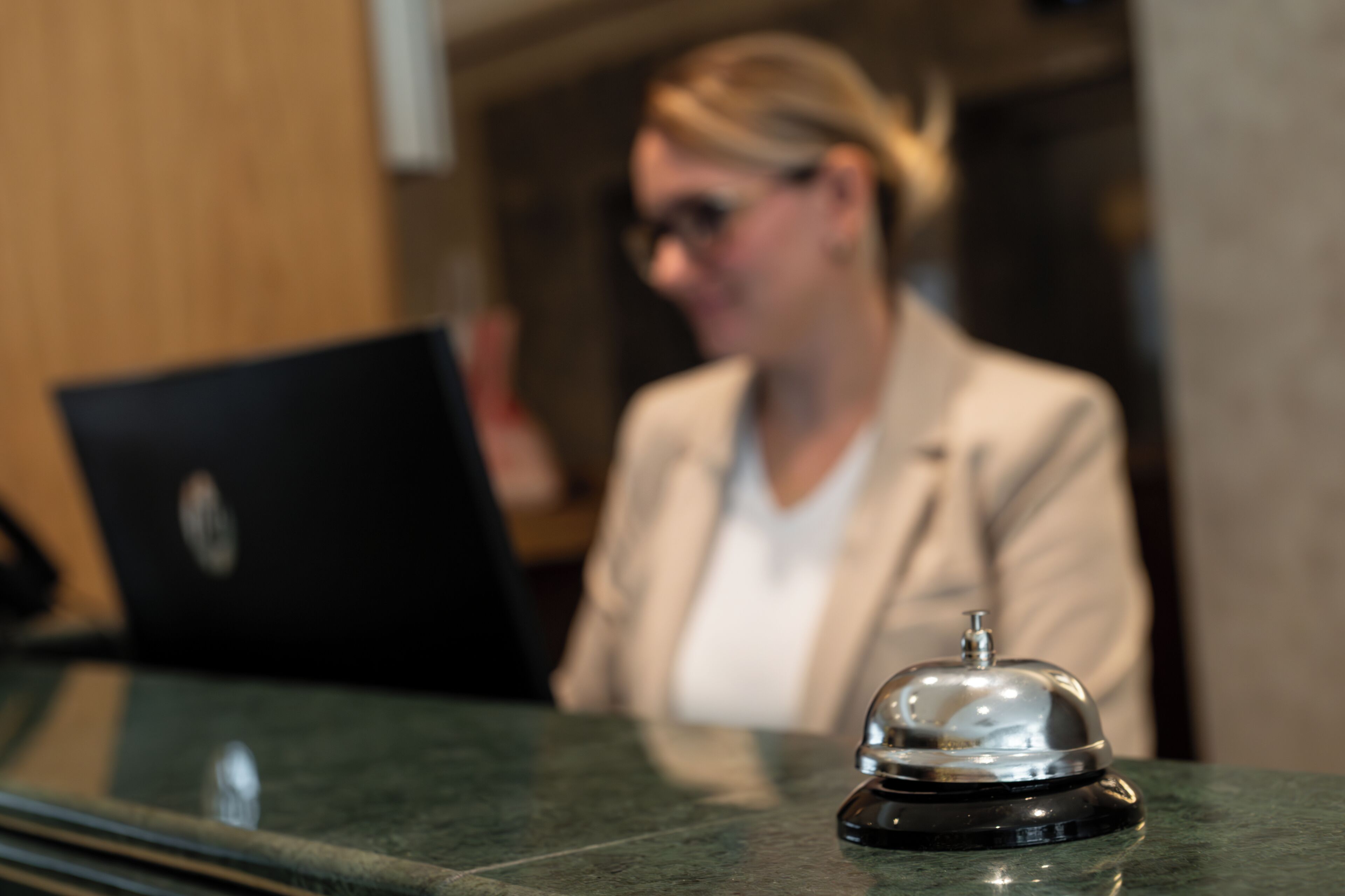 A reception desk with a black laptop, a silver bell, and a wooden panel in the background.