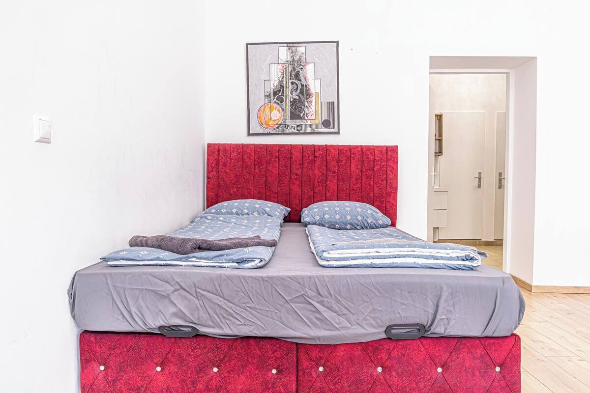 A bed with a red headboard, grey bedding, and a framed artwork above it.