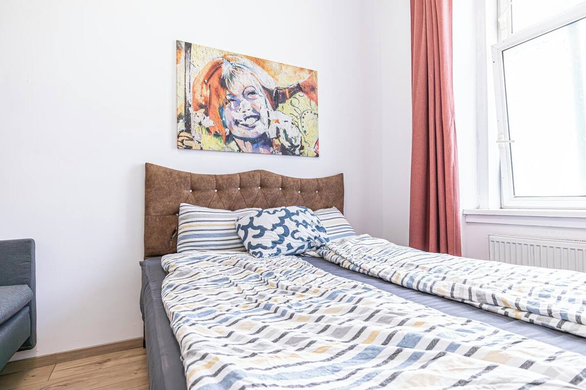 A bedroom with a bed, a gray chair, a colorful painting on the wall, and a window with curtains.