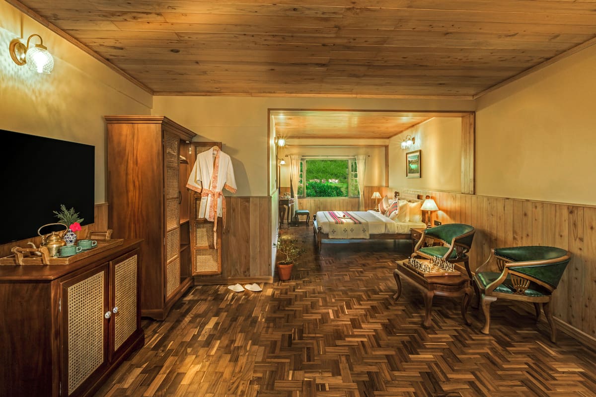 A room with a herringbone wooden floor, a bed with a colorful blanket, a desk with a lamp, and a view of a garden through an open door.