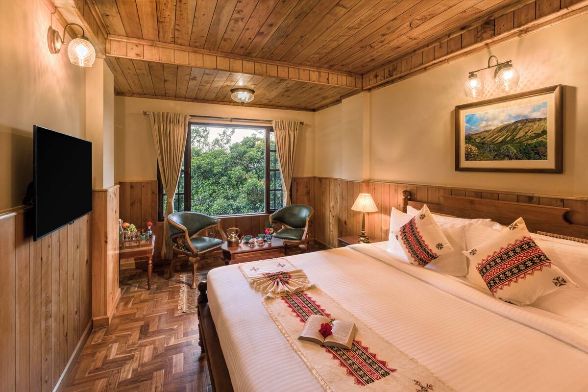 A hotel room with a wooden ceiling, a large bed with decorative pillows, a television, a dining table with chairs, and a window with curtains.
