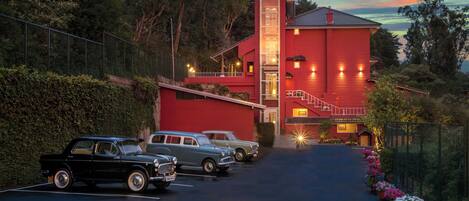 A red building with a staircase leading to a balcony, surrounded by greenery and parked vintage cars.