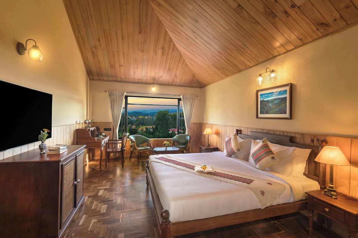 A bedroom with a wooden ceiling, a large bed with white linens, a television, a desk with a chair, and a view of the outdoors.