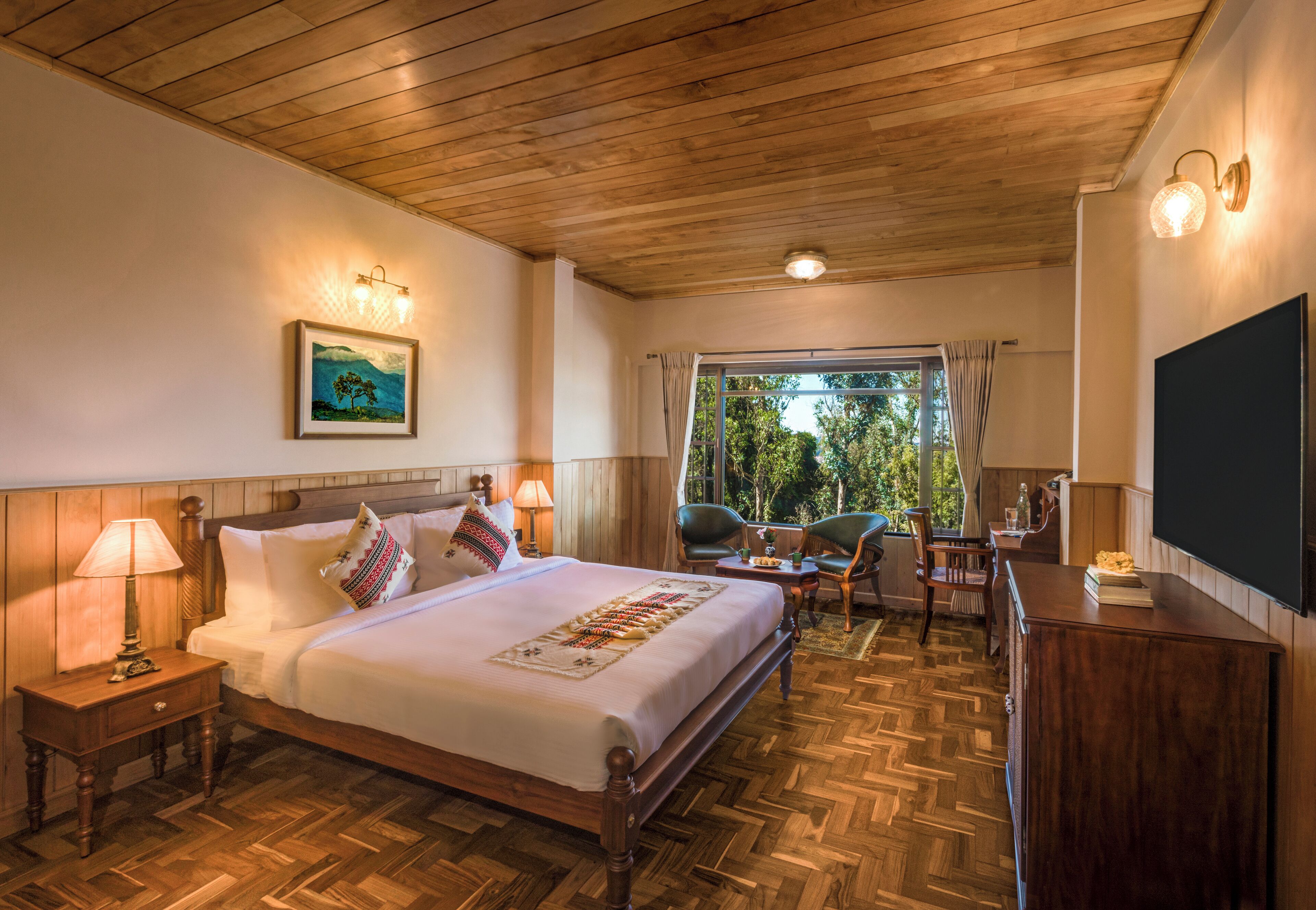 Quarto de hotel com teto de madeira, uma cama grande com colcha decorativa, duas mesas de cabeceira com luminárias, uma pequena mesa com cadeiras e uma janela grande com cortinas.