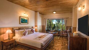 Une chambre d’hôtel avec un plafond en bois, un grand lit agrémenté d’une couvre-lit décorative, deux tables de chevet avec des lampes, une petite table avec des chaises et une grande fenêtre avec des rideaux.