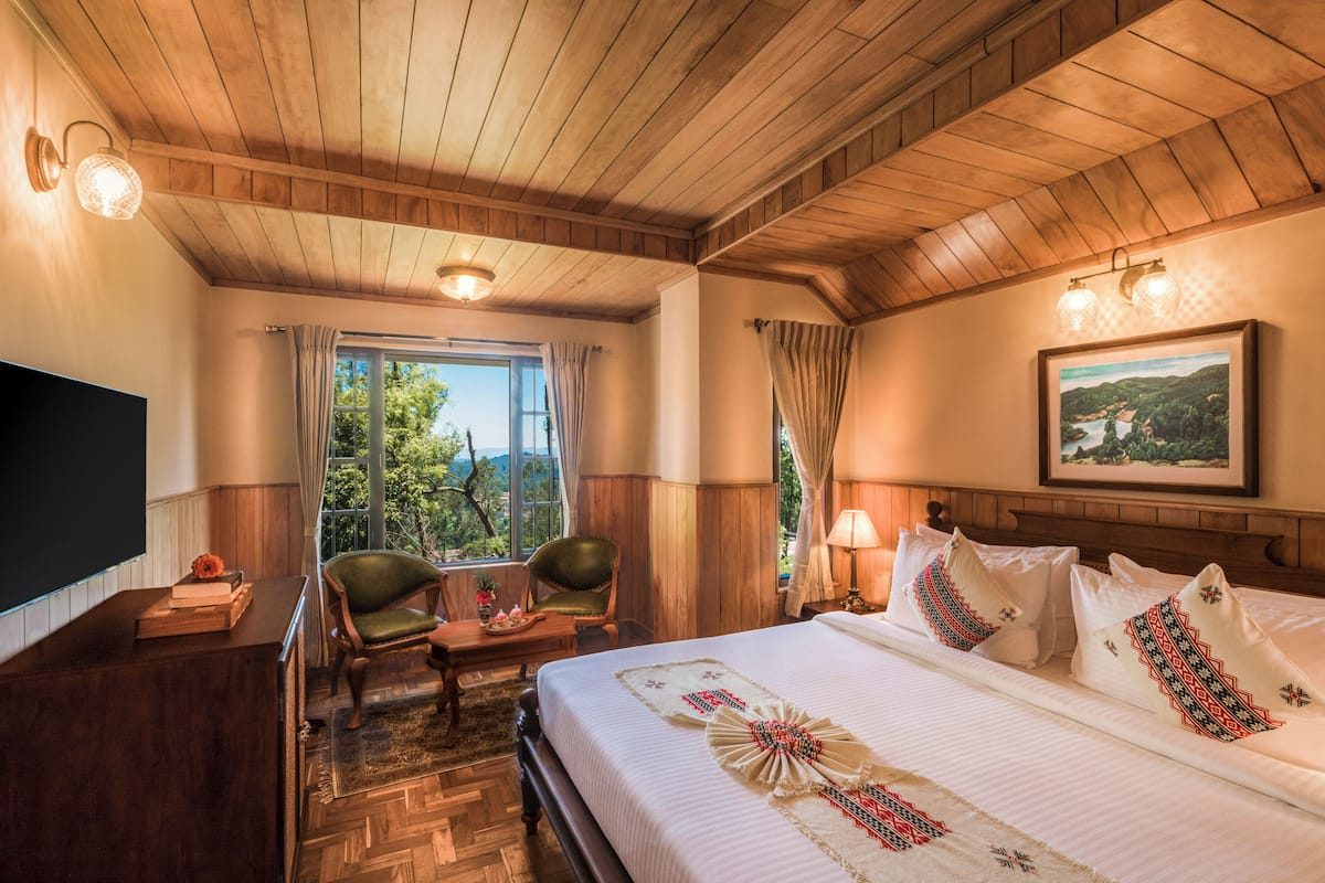 A hotel room with a wooden ceiling, a bed with decorative pillows, a desk with a flat-screen TV, and a chair by the window.