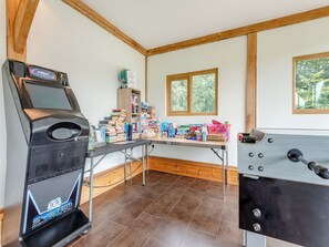 Una habitación con una consola de videojuegos, una mesa de madera con varios objetos y un ventanal con vistas a árboles.