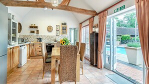 Une cuisine avec des meubles de rangement en bois, une table à manger avec des chaises en osier, un four encastré et une vue sur une piscine à travers de grandes fenêtres.