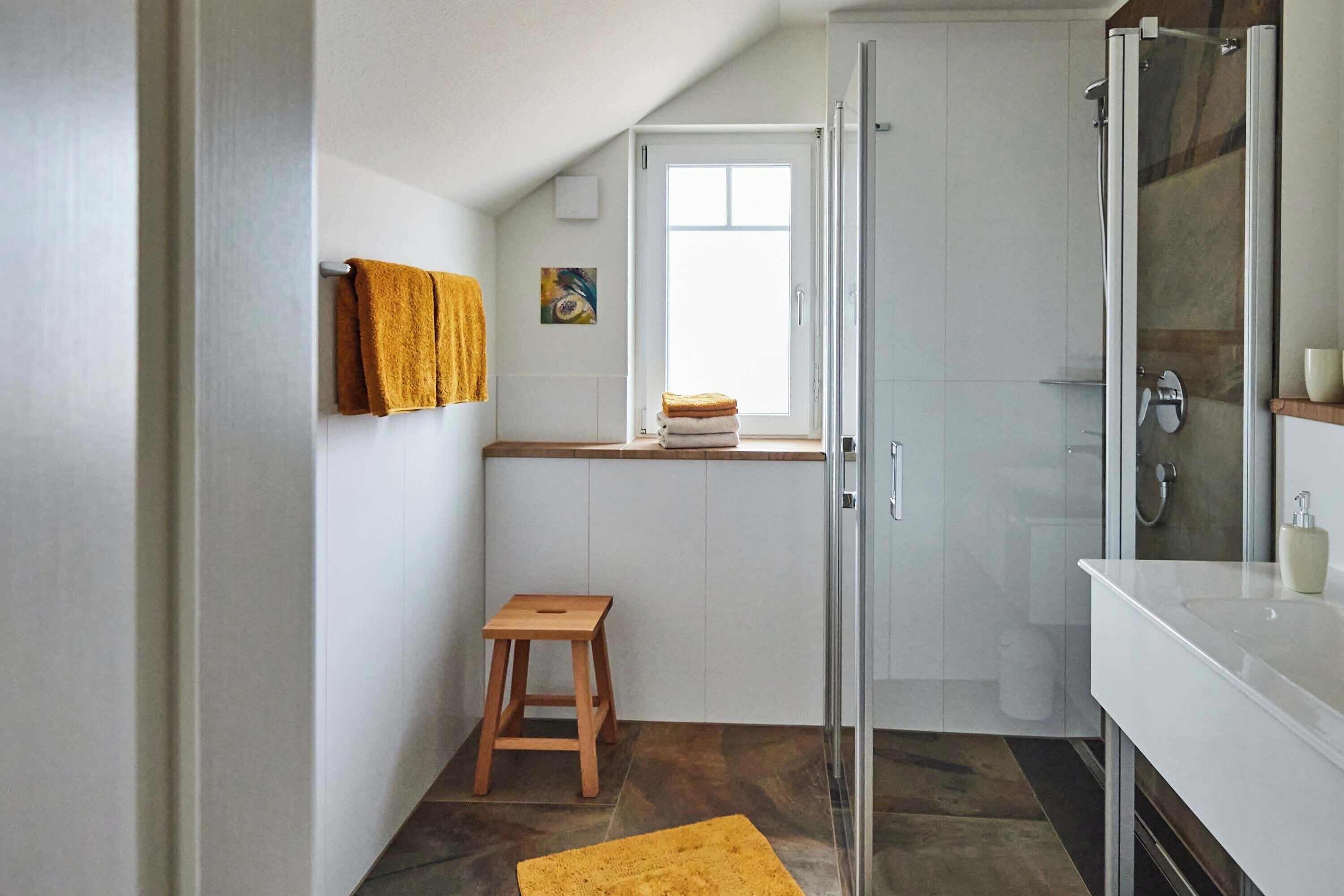 Une salle de bain moderne avec une douche à l’italienne en verre, un lavabo blanc et un tabouret en bois.