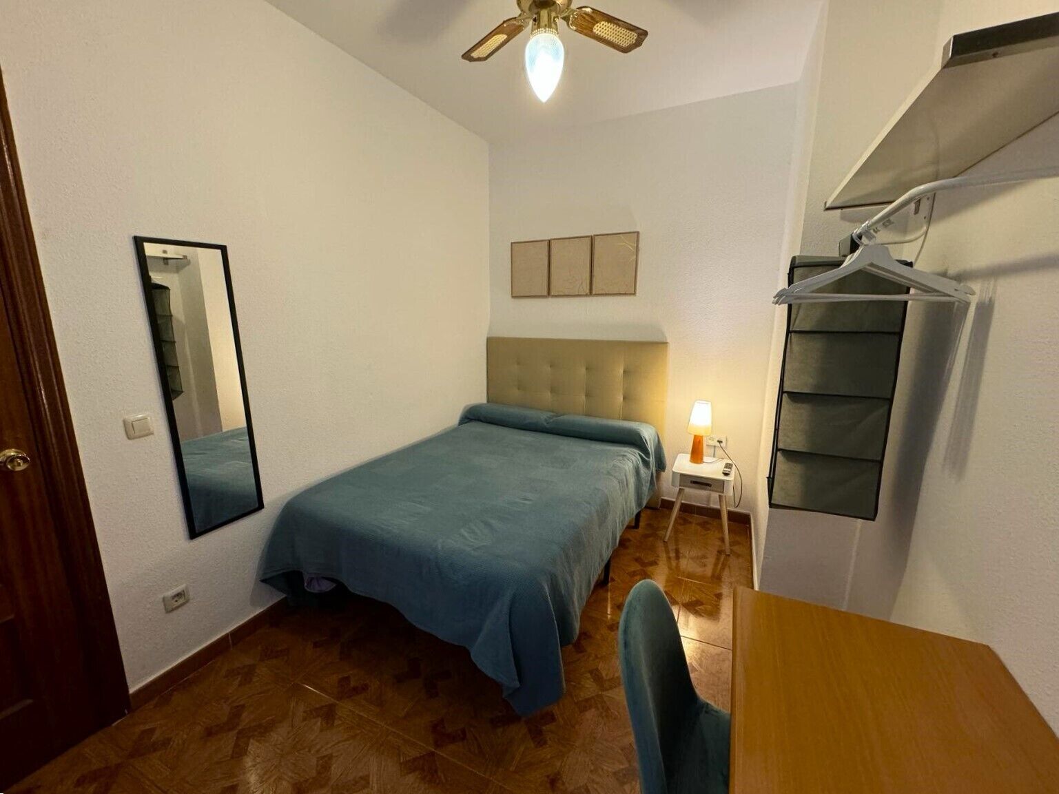 Une chambre à coucher comprenant un lit, une table de chevet avec une lampe, un miroir, un ventilateur de plafond et une armoire.