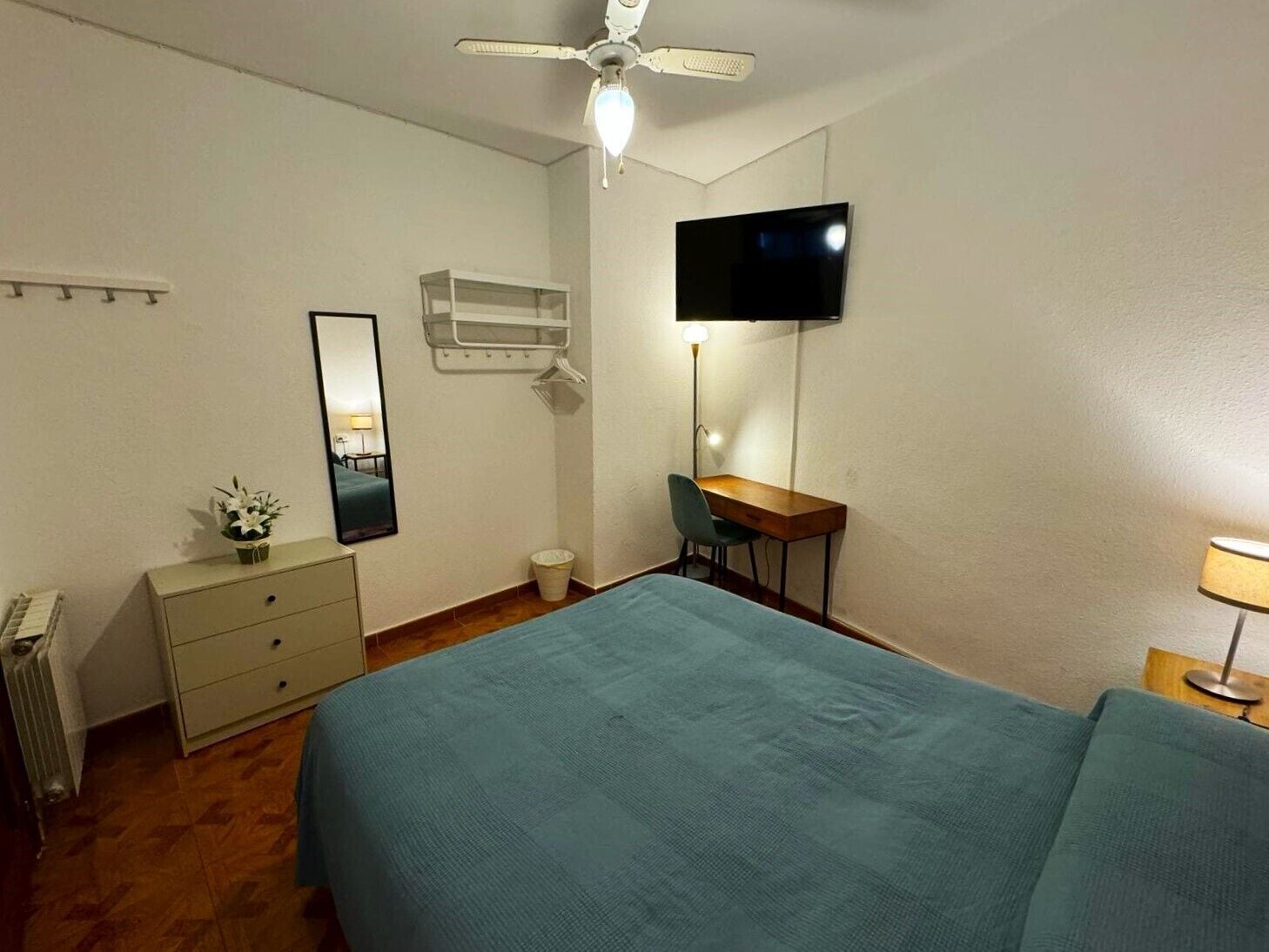 Een slaapkamer met een bed, een bureau, een stoel, een televisie, een spiegel, een radiator en een kleine plant.