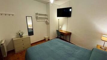 Een slaapkamer met een bed, een bureau, een stoel, een televisie, een spiegel, een radiator en een kleine plant.
