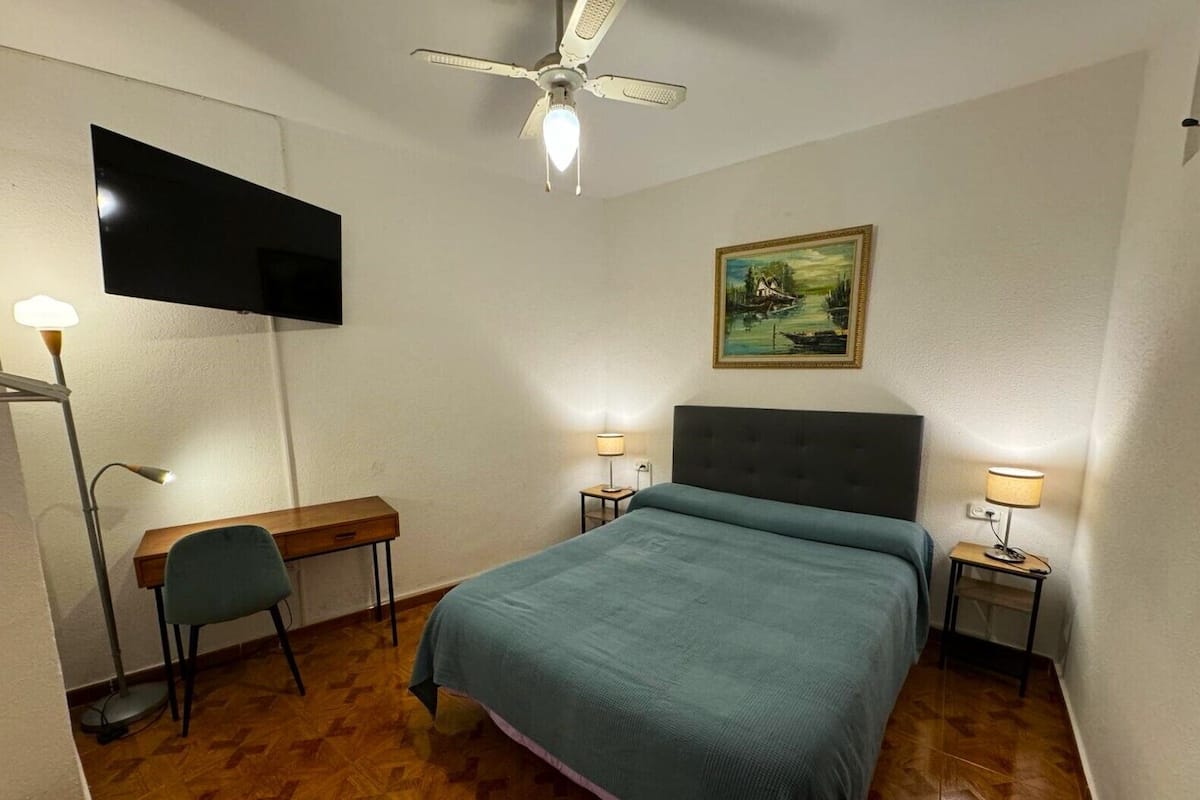 Een slaapkamer met een bed, een bureau, een stoel, twee bedlampjes, een plafondventilator en een schilderij aan de muur.